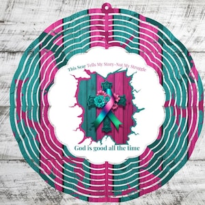 May include: A decorative wind spinner featuring a teal and pink color scheme. The design includes a cross with a ribbon and floral accents, set against a wood-grain background. Text reads "This Scar Tells My Story-Not My Struggle" and "God is good all the time."