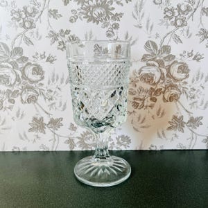 May include: A clear, vintage-style glass goblet with a diamond pattern on the bowl and a stemmed base. The glass is transparent, allowing light to pass through. The goblet is likely used for serving beverages.
