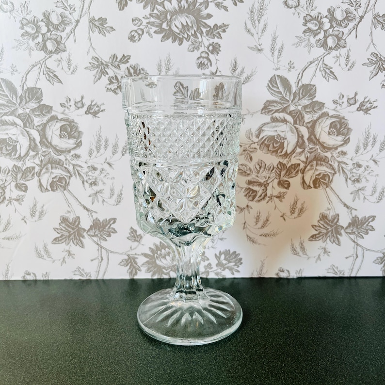 May include: A clear, vintage-style glass goblet with a diamond pattern on the bowl and a stemmed base. The glass is transparent, allowing light to pass through. The goblet is likely used for serving beverages.