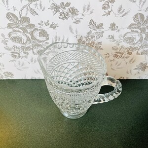 May include: A clear glass creamer with a diamond and textured pattern. The creamer has a handle and a spout. The background is a floral wallpaper.