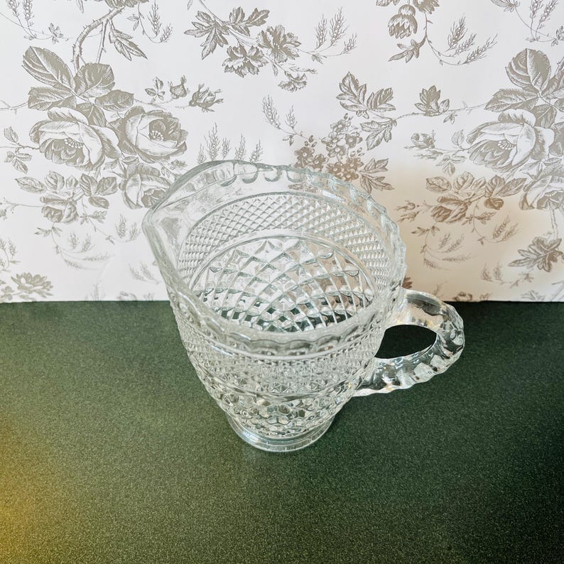 May include: A clear glass creamer with a diamond and textured pattern. The creamer has a handle and a spout. The background is a floral wallpaper.
