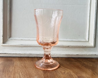 May include: A vintage pink glass goblet with a detailed floral and geometric pattern. The stemware has a rounded bowl and a pedestal base. The glass is a soft, translucent pink, and the goblet is set against a white background.