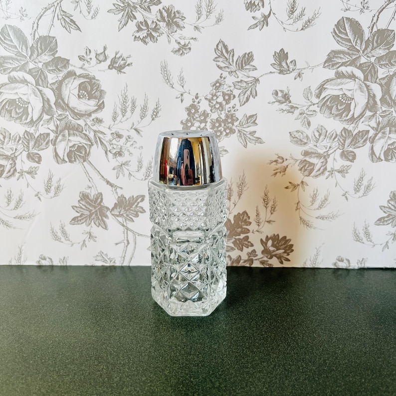 May include: A clear, faceted glass salt shaker with a shiny silver metal top. The salt shaker has a geometric pattern and is displayed against a floral wallpaper background. The shaker is a classic kitchen accessory.