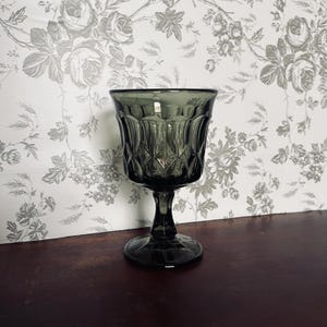 May include: A dark gray vintage-style goblet with a textured, diamond-patterned bowl and a stemmed base. The glass is set against a floral wallpaper background, creating a classic aesthetic. The goblet is likely made of glass.