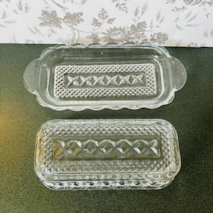 May include: Two clear glass butter dishes. The top dish has a scalloped edge and handles. Both dishes have a rectangular shape with a diamond and crosshatch pattern in the center. The dishes are on a dark green surface.