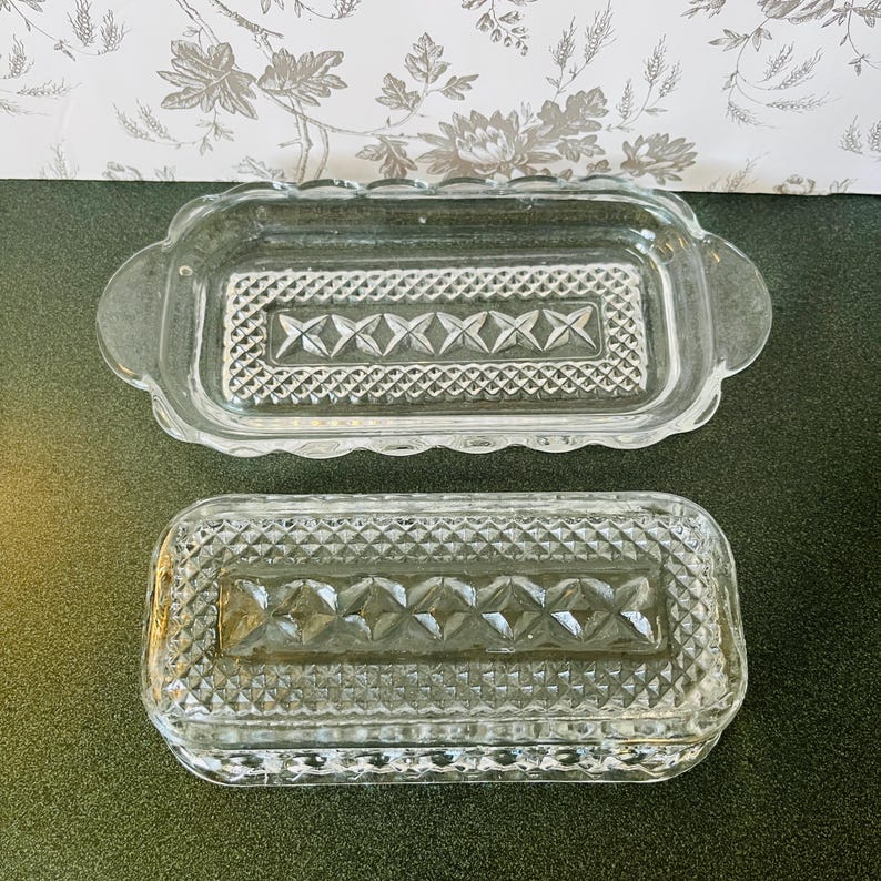 May include: Two clear glass butter dishes. The top dish has a scalloped edge and handles. Both dishes have a rectangular shape with a diamond and crosshatch pattern in the center. The dishes are on a dark green surface.