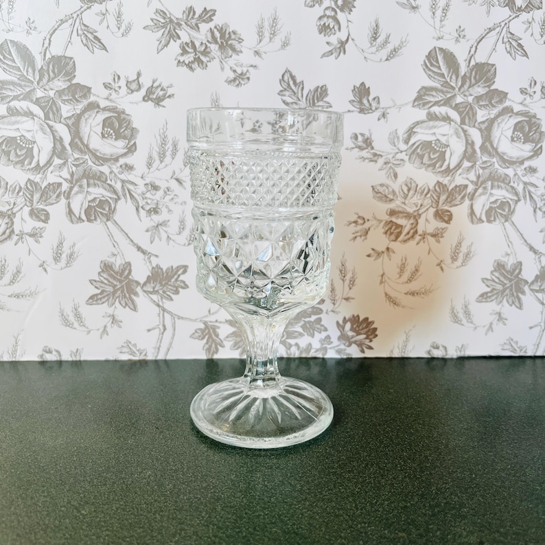 May include: A clear, vintage-style glass goblet with a diamond pattern on the bowl and a detailed stem. The goblet is made of clear glass and stands on a round base. The background features a floral wallpaper.