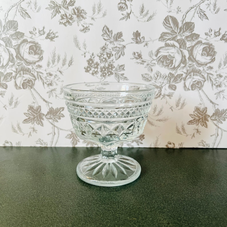 May include: A clear, vintage-style glass dessert dish with a decorative, diamond-patterned bowl and a stemmed base. The dish is made of clear glass and sits on a dark green surface, with a floral wallpaper background.
