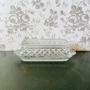 May include: A clear, rectangular glass butter dish with a lid. The dish features a diamond pattern and a scalloped edge. The background is a floral wallpaper.