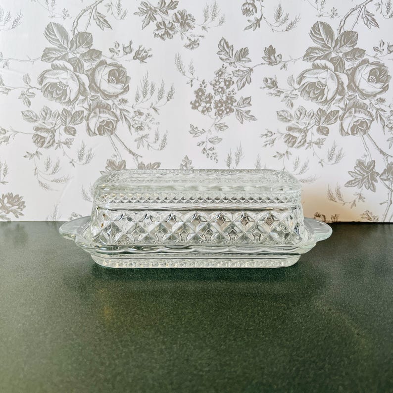 May include: A clear, rectangular glass butter dish with a lid. The dish features a diamond pattern and a scalloped edge. The background is a floral wallpaper.