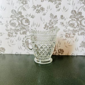 May include: A clear glass pitcher with a diamond pattern. The pitcher has a handle and a spout. The background is a floral wallpaper.