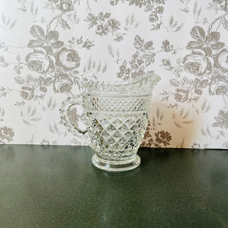May include: A clear glass pitcher with a diamond pattern. The pitcher has a handle and a spout. The background is a floral wallpaper.