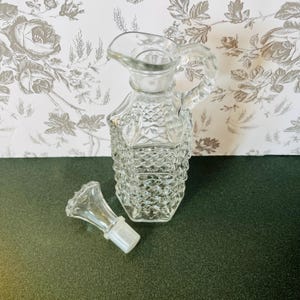 May include: A clear glass cruet with a handle and spout, featuring a diamond pattern on the body. The stopper is next to the cruet. The background is a floral wallpaper.