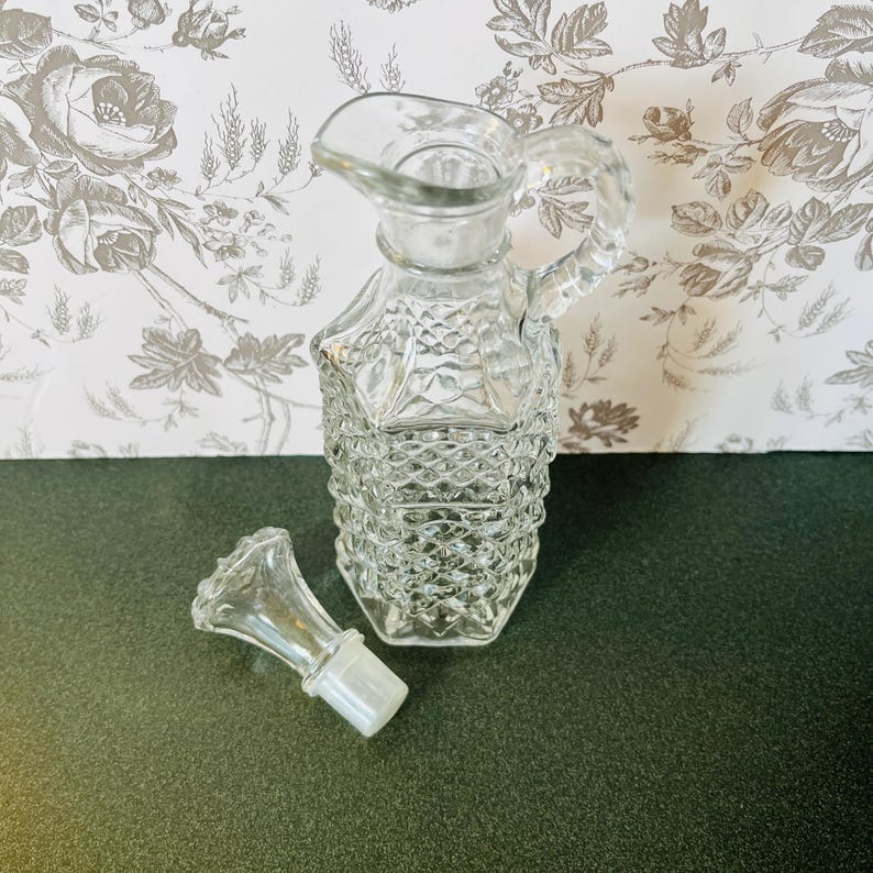 May include: A clear glass cruet with a handle and spout, featuring a diamond pattern on the body. The stopper is next to the cruet. The background is a floral wallpaper.