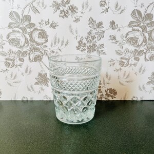 May include: A clear, vintage-style drinking glass with a diamond-cut pattern. The glass has a thick base and a cylindrical shape, perfect for serving beverages. The background features a floral wallpaper.