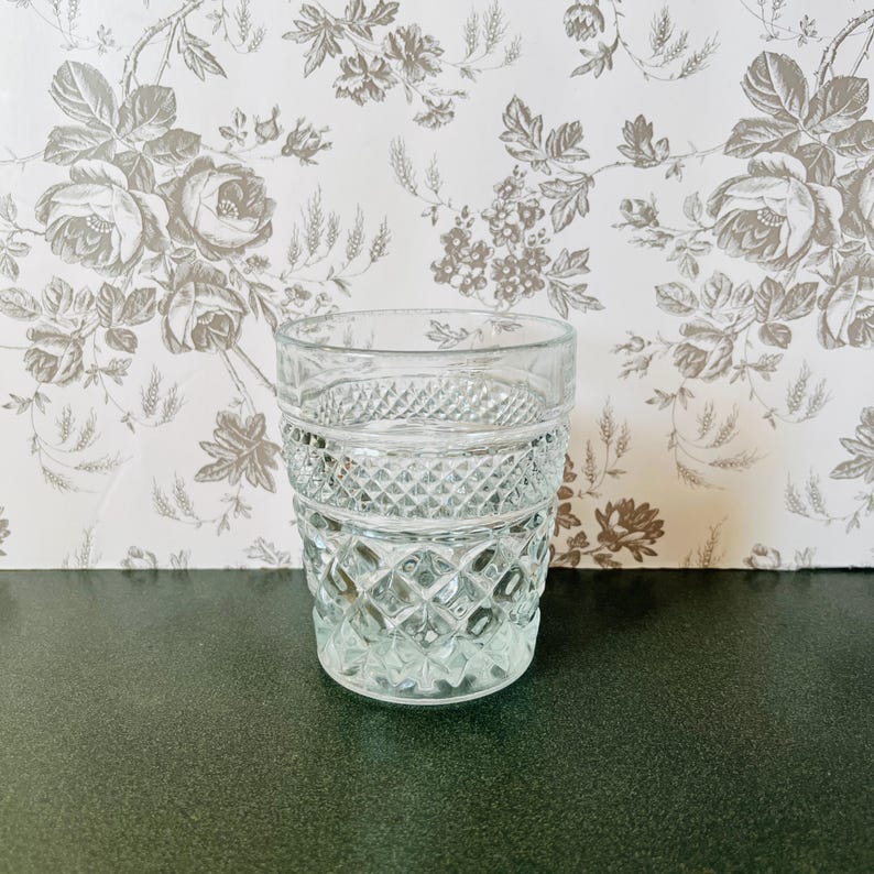 May include: A clear, vintage-style drinking glass with a diamond-cut pattern. The glass has a thick base and a cylindrical shape, perfect for serving beverages. The background features a floral wallpaper.