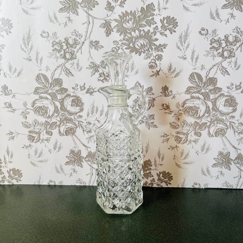 May include: A clear glass cruet with a diamond pattern, a handle, and a stopper. The cruet is on a dark surface against a floral wallpaper background. The cruet is designed for holding liquids like oil or vinegar.