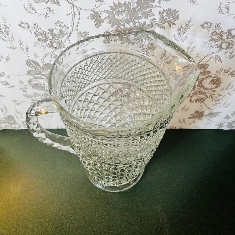 May include: A clear glass pitcher with a diamond pattern. The pitcher has a handle and a spout for pouring. The pitcher is likely used for serving beverages. The background features a floral pattern.
