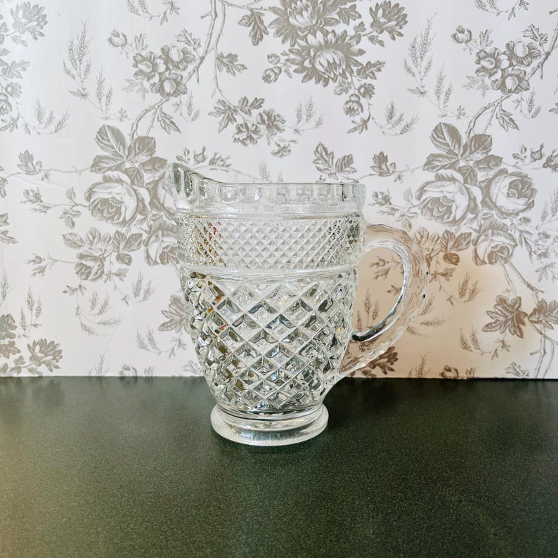 May include: A clear glass pitcher with a diamond pattern. The pitcher has a handle and a spout for pouring. The background features a floral wallpaper.