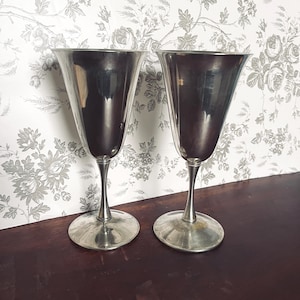 May include: Two silver-colored wine glasses with a flared bowl and slender stem. The glasses sit on a round base. The background is a floral wallpaper in shades of gray and white.