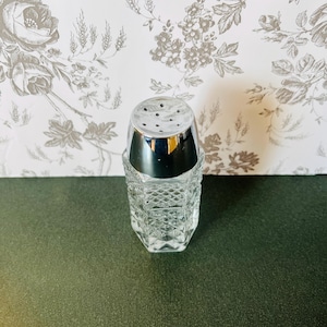 May include: A clear glass salt shaker with a chrome top. The shaker has a diamond pattern and is sitting on a dark green surface. The background is a floral wallpaper.