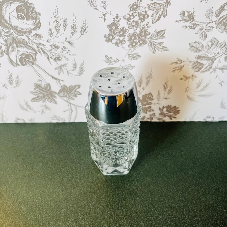May include: A clear glass salt shaker with a chrome top. The shaker has a diamond pattern and is sitting on a dark green surface. The background is a floral wallpaper.