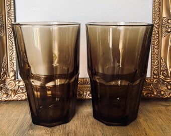 May include: Two brown-tinted drinking glasses with a faceted design. The glasses have a rectangular shape with a hexagonal base. The glasses are made of a transparent material, allowing light to pass through. The glasses are set against a wooden surface.