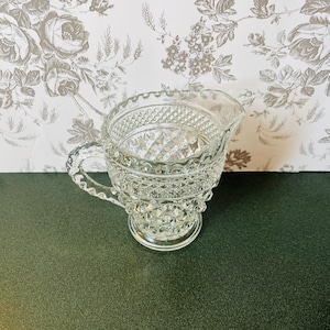 May include: A clear glass creamer with a diamond pattern and a handle. The creamer has a scalloped rim and sits on a round base. The background features a floral wallpaper.