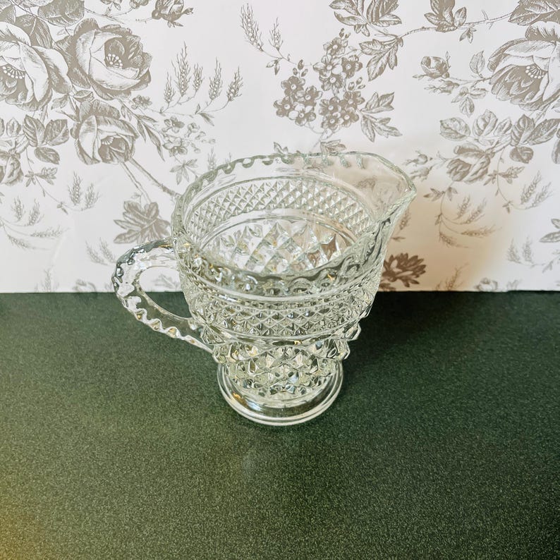 May include: A clear glass creamer with a diamond pattern and a handle. The creamer has a scalloped rim and sits on a round base. The background features a floral wallpaper.