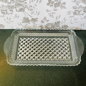 May include: A rectangular clear glass serving tray with a diamond pattern in the center and a scalloped edge. The tray has two rounded handles on either side. The background features a floral wallpaper.