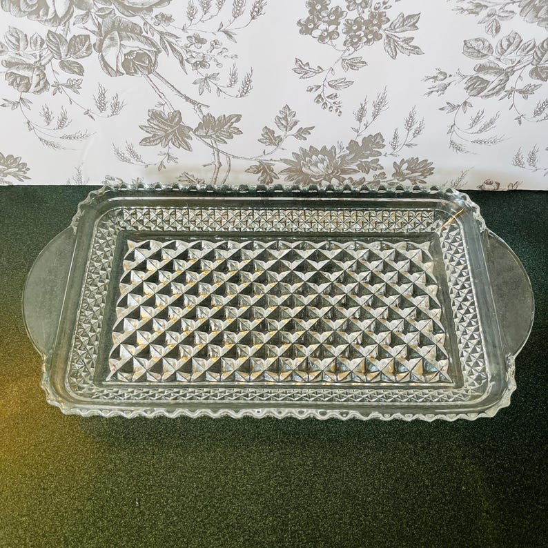 May include: A rectangular clear glass serving tray with a diamond pattern in the center and a scalloped edge. The tray has two rounded handles on either side. The background features a floral wallpaper.