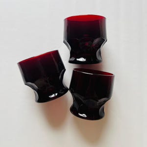 May include: Three ruby red drinking glasses with a unique, faceted design. The glasses have a short, squat shape and a deep, rich color. The glasses are made of a thick glass material.