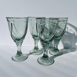 May include: Four vintage green-tinted wine glasses. Each glass features a decorative leaf design on the bowl and stem. The glasses are arranged on a white surface, with shadows adding depth to the composition. The glasses are made of glass.