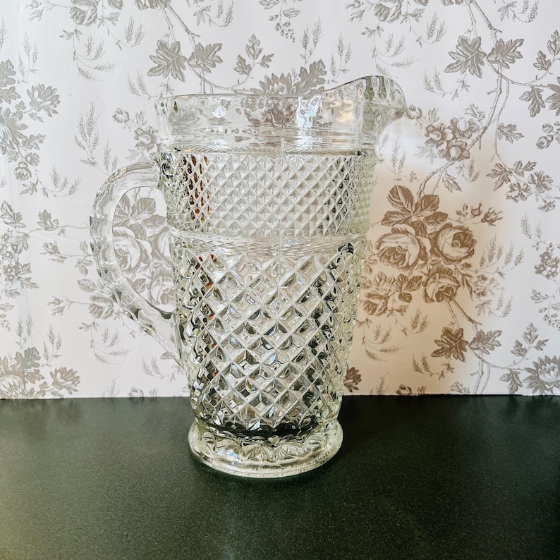 May include: Clear glass pitcher with a diamond pattern. The pitcher has a handle and a spout for pouring. The design is repeated throughout the body of the pitcher. The pitcher is sitting on a dark surface.