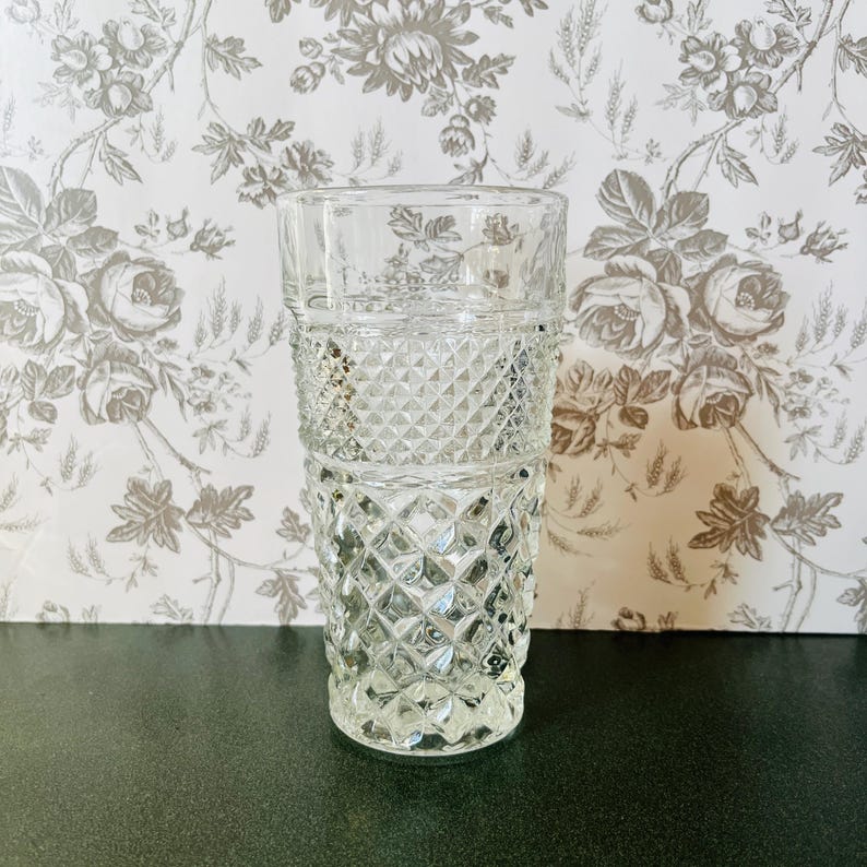 May include: A clear, vintage-style drinking glass with a diamond-patterned design. The glass is cylindrical with a thick base and a slightly flared rim. The glass is made of clear glass and is likely used for water or other beverages.