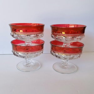 May include: Four vintage clear glass dessert bowls with a ruby red rim and base. The bowls have a decorative pattern around the middle and a clear stem and base. The bowls are likely from the mid-century.