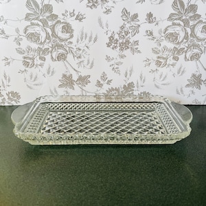 May include: A clear glass rectangular serving tray with a diamond pattern in the center and a decorative border. The tray has rounded handles on each end. The background features a floral wallpaper.