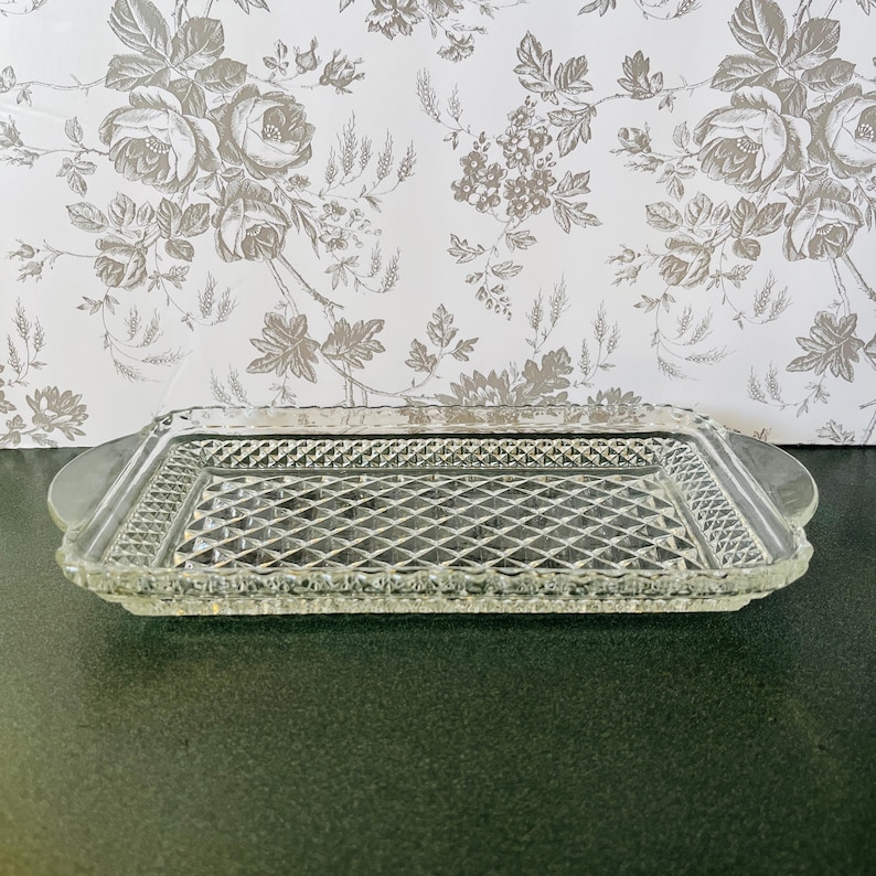 May include: A clear glass rectangular serving tray with a diamond pattern in the center and a decorative border. The tray has rounded handles on each end. The background features a floral wallpaper.