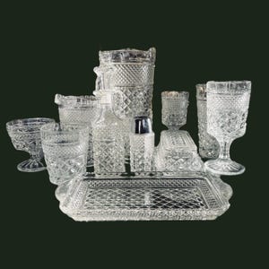 May include: A collection of clear, diamond-patterned glassware, including a pitcher, glasses, a tray, and a sugar bowl. The set is arranged on a dark green background, showcasing the intricate details of each piece. The glassware appears to be vintage.