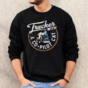 Trucker with a copilot cat grunge style round design on Gildan 18000 sweatshirt.  Calico cat wearing a trucker cap sitting on a big rig 18-wheeler truck.  Male model wearing black sweatshirt.