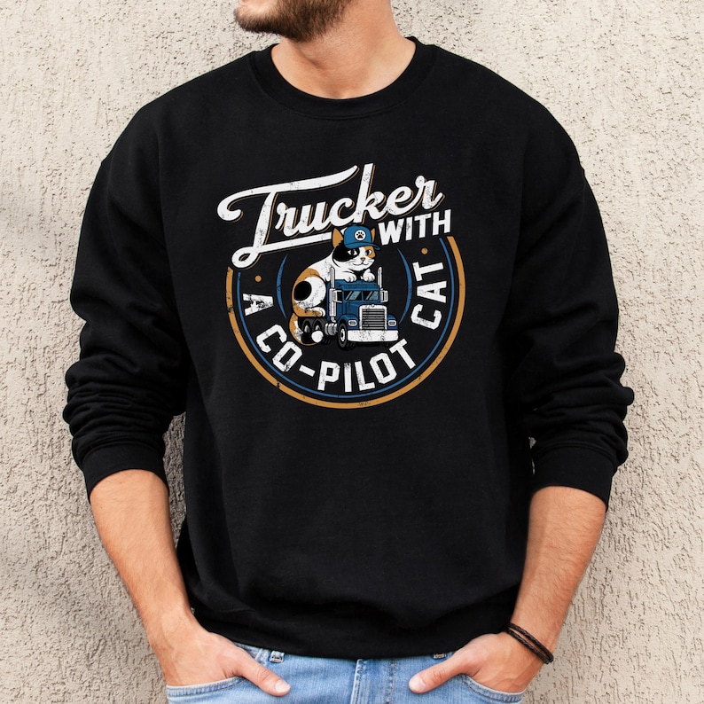 Trucker with a copilot cat grunge style round design on Gildan 18000 sweatshirt.  Calico cat wearing a trucker cap sitting on a big rig 18-wheeler truck.  Male model wearing black sweatshirt.