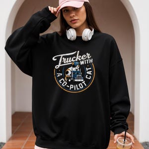 Trucker with a copilot cat grunge style round design on Gildan 18000 sweatshirt.  Calico cat wearing a trucker cap sitting on a big rig 18-wheeler truck.  Female model wearing black sweatshirt.