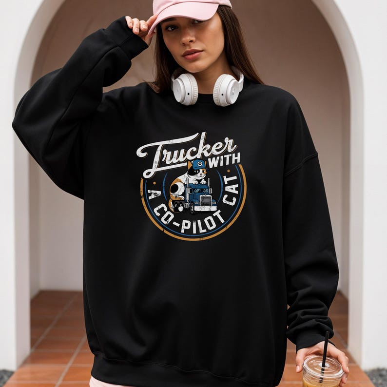 Trucker with a copilot cat grunge style round design on Gildan 18000 sweatshirt.  Calico cat wearing a trucker cap sitting on a big rig 18-wheeler truck.  Female model wearing black sweatshirt.