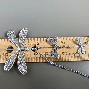 May include: Silver necklace with three dragonfly charms. The largest dragonfly is 2 inches long and the necklace is 24 inches long.