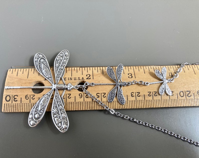 May include: Silver necklace with three dragonfly charms. The largest dragonfly is 2 inches long and the necklace is 24 inches long.