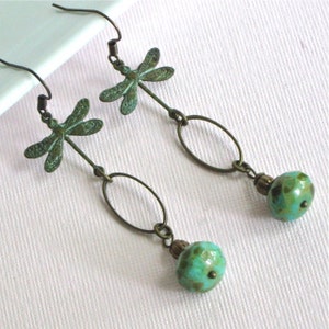 May include: A pair of dangle earrings with a dragonfly charm and a turquoise bead. The dragonfly is made of metal and has a green patina. The bead is round and has a speckled pattern.