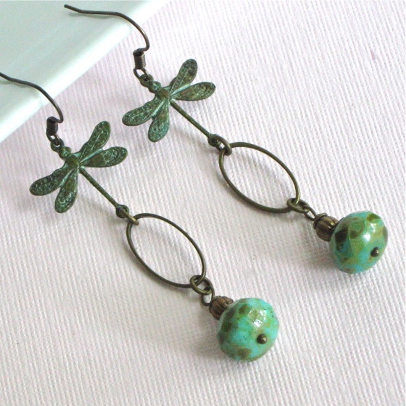 May include: A pair of dangle earrings with a dragonfly charm and a turquoise bead. The dragonfly is made of metal and has a green patina. The bead is round and has a speckled pattern.