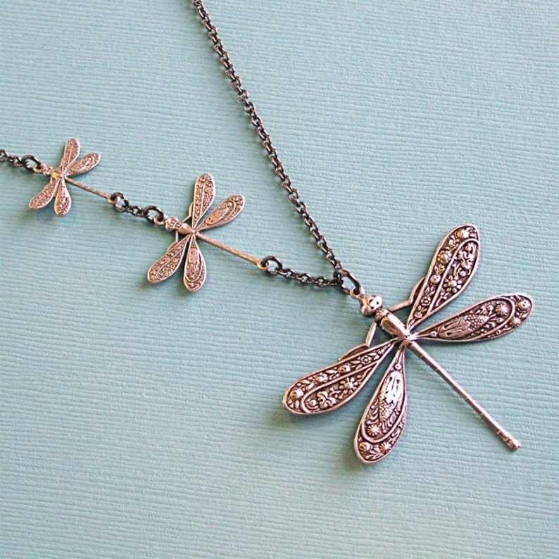 May include: A silver necklace with three dragonfly charms. The dragonflies are detailed with intricate patterns and have a vintage look.
