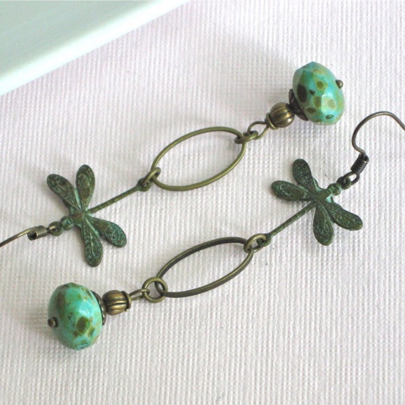 May include: A pair of dangle earrings with turquoise and bronze accents. Each earring features a dragonfly charm, a bronze oval link, and a turquoise bead.