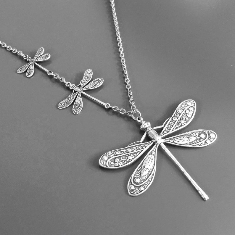 May include: Silver necklace with three dragonfly charms. The largest dragonfly charm is at the bottom of the necklace and the two smaller charms are above it.